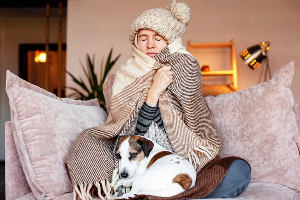 woman wrapped in a blanket with a beanie on her head while she sits on the couch inside because it's cold